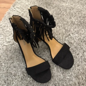 Svarta sandaler med fransar och klack - Svarta sandaler med öppen tå, bred rem över tårna och chunky klack i trälook. Runt ankeln finns snygga fransdetaljer och små guldfärgade nitar. Skorna är i mockaimitation och har dragkedja bak för enkel påtagning. Perfekta för dig som vill sticka ut lite extra.