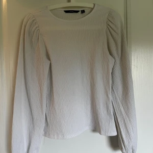 Vit långärmad blus från Vero Moda - Säljer en stilren vit blus från Vero Moda i storlek S. Blusen har ett strukturerat tyg med lätt rynkade axlar och långa ärmar. Rund halsringning och rak passform gör den enkel att matcha med olika outfits. Perfekt för dig som gillar minimalistisk stil.