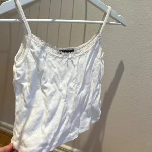 Vit kort spetsdetaljerad linnetopp  - Supersöt kort vit linnetopp från Brandy Melville med tunna axelband, spetskant och liten rosett framtill. Toppen har diskret hålmönster i tyget och är perfekt för en fräsch och enkel look. Materialet är mjukt och bekvämt.