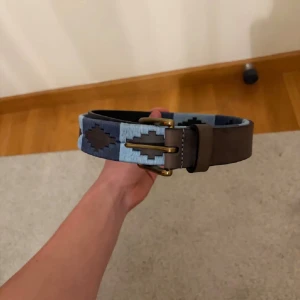 Blått och brunt mönstrat bälte - Säljer ett snyggt bälte med geometriskt mönster i olika blå nyanser och brunt. Bältet har en robust metallspänne och är tillverkat i skinn med textildetaljer. Perfekt för att ge outfiten lite extra edge. priset går att diskutera.
