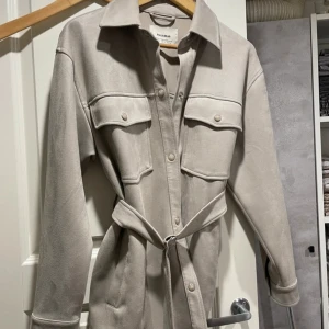 Grå jacka  från Pull&Bear - Stilren grå jacka från Pull&Bear i mjuk mockaimitation. Jackan har långa ärmar, stora bröstfickor med knappar och ett matchande bälte i midjan. Perfekt för dig som gillar minimalistisk och trendig look.