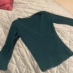 Mörkgrön ribbad omlottopp från Bershka - Snygg mörkgrön omlottopp från Bershka i ribbad bomull. Toppen har halv lång ärm och en djup v-ringning som ger en cool och modern look. Perfekt att styla med jeans eller kjol för en trendig vibe.