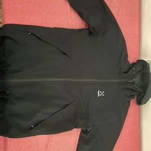 Haglöfs jacka - Svart vindjacka från Haglöfs med snyggt broderad logga på bröstet. Jackan har huva med snörning, två sidofickor med dragkedja och meshfoder på insidan. Perfekt för blåsiga dagar och outdoor-aktiviteter. Stilren och funktionell design.