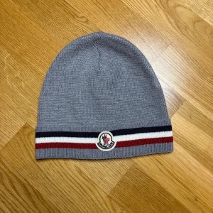 Grå Moncler mössa  - Snygg grå mössa från Moncler med klassiska ribbade ränder i svart, vitt och rött längst ner. Mössan är stickad i 100% ull och har Moncler-logga framtill. Perfekt för dig som vill ha en clean och stilren look med lite lyxig känsla.
