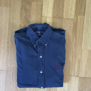 Blå skjorta från Ralph Lauren - Snygg blå skjorta från Ralph Lauren med klassisk krage och knappar framtill. Skjortan har en röd broderad logga på bröstet och är tillverkad i mjuk bomull. Perfekt för dig som gillar stilrena och tidlösa plagg.
