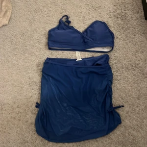 Mörkblå bikini med kjol och omlott - Supersnygg mörkblå bikini med omlott-topp och matchande höga bikinitrosor. Setet kommer med en meshkjol med snörning på sidorna för extra stilpoäng. Bikinin har dubbla axelband och är tillverkad i mjukt syntetmaterial som torkar snabbt. Perfekt för strandhäng och poolparty!