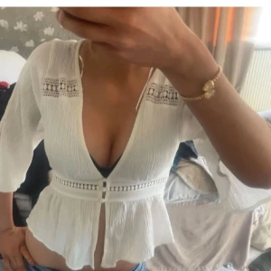 Vit blus med spets och v-ringning - Säljer en vit blus med djup v-ringning och fina spetsdetaljer vid axlarna. Blusen har trekvartsärm och ett markerat midjeband med dekorativa hål. Materialet är lätt och luftigt, perfekt för vår och sommar. Snygg och feminin stil!