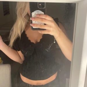 Svart stickad blus med volanger - Säljer en svart stickad blus med volangdetaljer och v-ringad hals. Toppen har korta ärmar och ett genombrutet mönster 