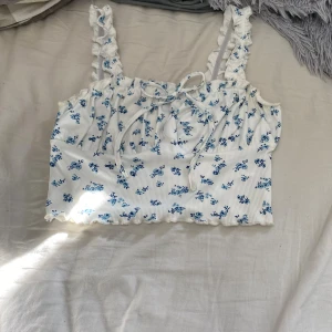 Vit blommig linnetopp med volangband - Supersöt vit linnetopp med blått blommönster, volangprydda axelband och liten knytning framtill. Toppen är croppad med vågig kant nertill och har en romantisk vibe. Säljs då den är för stor, knappt använd.