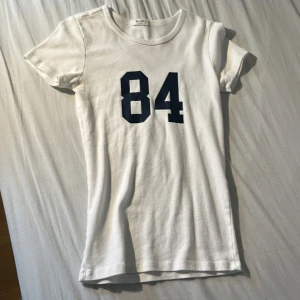 Vit t-shirt med tryck köpt från Brandy Melville  - Vit t-shirt från Brandy Melville med mörkblått tryck av siffran 84 på bröstet. Klassisk rund halsringning och korta ärmar. Gjord i mjuk bomull för en skön känsla. Perfekt för en avslappnad och sportig stil. Observera att siffrorna är lite slitna 100% bomull 