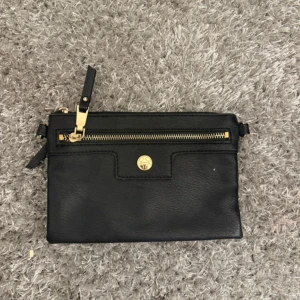 Svart kuvertväska med gulddetaljer - Snygg svart kuvertväska i skinn med guldfärgad dragkedja och logoknapp framtill. Perfekt storlek för det viktigaste och stilren design som passar till många outfits. Skriv för fler bilder 