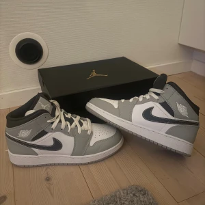 Nike Air Jordan 1 Mid grå/vit - Nike Air Jordan 1 Mid sneakers i grått och vitt med svarta detaljer och klassisk Swoosh på sidan. Skorna har snörning, rund tå och platt sula. Materialet är skinn och syntet, och de kommer med originalkartong med guldfärgad Jumpman-logga.