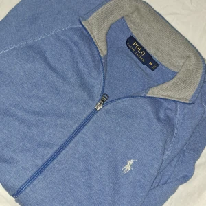 Blå ziptröja från Polo Ralph Lauren - Snygg blå ziptröja från Polo Ralph Lauren med grå ribbad krage och klassisk broderad logga på bröstet. Tröjan har hel dragkedja och är perfekt för lager-på-lager-stil. Skön och stilren, passar till allt.