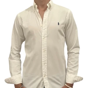 Skjorta - Ralph Lauren - Klassisk vit skjorta från Ralph Lauren med blå broderad logga på bröstet. Skjortan har lång ärm, knappar hela vägen och är tillverkad i 100% bomull. Snygg passform och tidlös stil som funkar till allt. Storlek 20 - motsvarar S