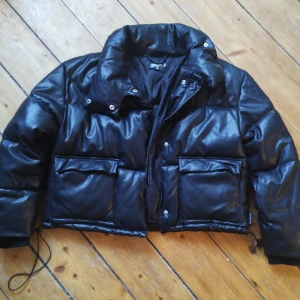 Svart pufferjacka i skinnlook Missguided stl 38 - Säljer en svart pufferjacka från Missguided Petite i storlek 38. Jackan har en glansig skinnlook, stora fickor framtill, hög krage och dragsko nertill. Stängs med dragkedja och tryckknappar. Perfekt för kalla dagar när du vill ha en edgy vibe.