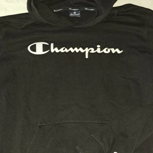 Svart Champion hoodie med logga - Svart hoodie från Champion i storlek L med stor vit broderad logga över bröstet. Klassisk huva och känguruficka framtill. Perfekt för en chill och sportig stil. Mjuk och skön bomullsblandning.