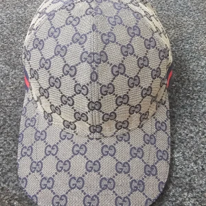 Gucci beige keps med GG-mönster - Snygg beige keps från Gucci med klassiskt GG-mönster i mörkblått. Kepsen har en böjd skärm och en mörkblå och röd rand på sidan. Tillverkad i bomull med detaljer i skinn och justerbar rem baktill. Perfekt för dig som vill ha en lyxig streetstyle-look.