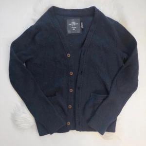 Mörkblå kofta från H&M L.O.G.G. - Mysig mörkblå kofta från H&M L.O.G.G. med v-ringning, ribbade muddar och bruna knappar framtill. Koftan har två fickor och är stickad i 80% ull och 20% polyamid, perfekt för kyliga dagar.