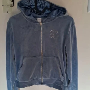 Blå velour hoodie och byxor - Mjuk och skön blå hoodie i velour från Linda L med glittrande strassdetalj på bröstet. Tröjan har huva, dragkedja framtill och praktisk ficka. Ärmarna är långa och nederkanten har snörning för extra stilpoäng.