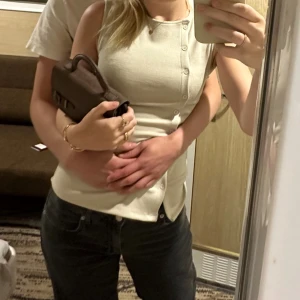 Beige ärmlös linnetopp med knappar - Säljer en stilren beige linnetopp med rund hals och knappar längs framsidan. Toppen är ärmlös och har en slim passform som sitter snyggt mot kroppen. Perfekt för dig som gillar minimalistisk och clean stil.