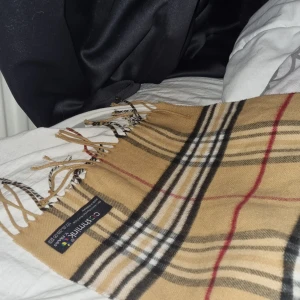 Beige rutig halsduk från Cashmink - Klassisk halsduk i beige med rutigt mönster i svart, vitt och rött från Cashmink. Mjuk och lätt, tillverkad i 100% akryl. Avslutas med fransar i ändarna för en snygg touch. Perfekt accessoar för att lyfta din outfit.