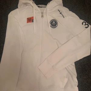 Limited Edition kofta Sail racing - Limited edition vit hoodie med dragkedja och huva från Sail racing. Snygga tryck på både fram- och baksidan, samt nummerdetalj på ärmen. Tröjan har snörning i huvan och praktiska fickor framtill. Mindre i storlek , Strl L men passar M