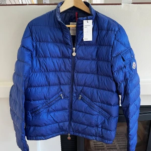 Moncler jacka herr blå - Snygg blå pufferjacka från Moncler med klassisk logga på ärmen, två dragkedjefickor framtill och quiltad design. Jackan har hög krage, lång ärm och stängs med dragkedja. Perfekt för dig som vill ha en stilren och sportig look. ÄKTA Size 4-L