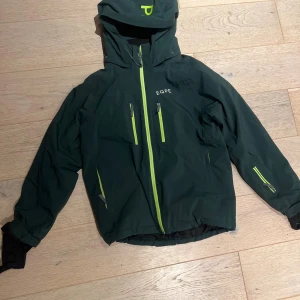 Mörkgrön viterjacka från EQPE - Snygg mörkgrön vindjacka från EQPE med huva och limegröna dragkedjor. Jackan har flera praktiska fickor med dragkedja och en cool logga på bröstet. Perfekt för dig som gillar att vara ute och vill ha en jacka med sportig vibe. Skidjacka
