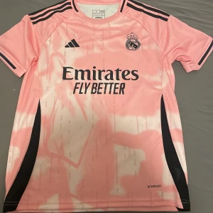 Real Madrid rosa fotbollströja Adidas S - Snygg Real Madrid fotbollströja från Adidas i slim fit, storlek S. Tröjan är rosa med svarta detaljer och har ett abstrakt mönster på både fram- och baksidan. Klassisk rund hals, korta ärmar och klubbmärke på bröstet. Tillverkad i lätt och ventilerande polyester.