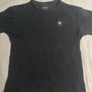 Svart t-shirt från Lyle & Scott - Svart t-shirt från Lyle & Scott Junior med klassisk rund hals och korta ärmar. På bröstet sitter en svart patch med den ikoniska gula örnen. T-shirten är gjord i mjuk bomull och har en clean, stilren look som funkar till allt.