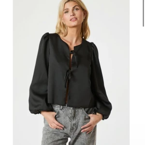 Svart satinblus från Neo Noir - Supersnygg svart blus i satin från Neo Noir med puffiga långärmar och två knytband framtill. Helt ny, lappen kvar! Köpt för 749kr