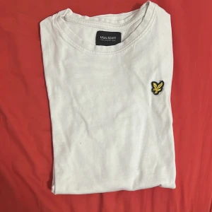 Vit t-shirt från Lyle & Scott - Vit t-shirt från Lyle & Scott med klassisk rund hals och ikonisk gul logga broderad på bröstet. Enkel och stilren design i mjuk bomull, perfekt till jeans eller shorts. Passar dig som gillar en clean och tidlös look.