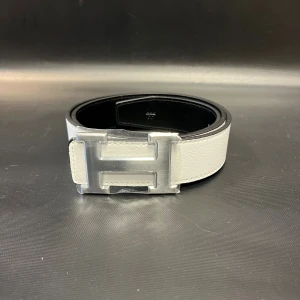 H logga bälte vändbart svart/vitt skärp - Snyggt vändbart H logga bälte i svart och vitt skinn med en stor, ikonisk H-spänne i silverfärgad metall. Bältet har en stilren och modern look med slät yta och tydliga sömmar. Perfekt för att lyfta din outfit med en exklusiv touch.