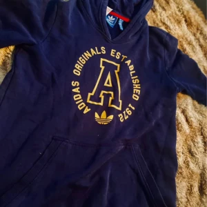 Mörkblå Adidas hoodie med gul print - Säljer en mörkblå hoodie från Adidas Originals med stor gul A och texten 'Adidas Originals Established 1949' på bröstet. Klassisk känguruficka, huva och mjukt material. Perfekt för dig som gillar sportig och avslappnad stil.