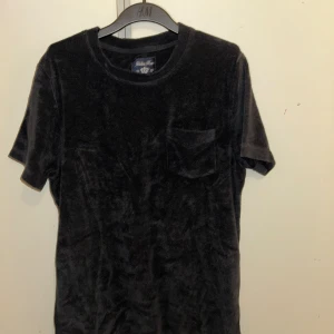 Svart sammetst-shirt Robbie Moor - Svart t-shirt i mjuk sammet från Robbie Moor. Klassisk rund halsringning, korta ärmar och en liten bröstficka. Perfekt för dig som gillar en stilren look med lite extra känsla tack vare materialet.