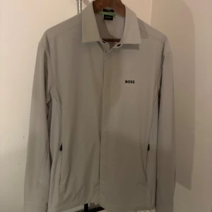 Beige overshirt från BOSS - Stilren beige overshirt från BOSS med klassisk krage och svarta knappar. Skjortan har diskret BOSS-logga på bröstet och två dragkedjeförsedda fickor i sidorna. Perfekt för lager-på-lager och ger en clean look. Han på bilden är 180 cm lång. Medium i Storlek derav för stor. Perfekt för 185 cm och uppåt!