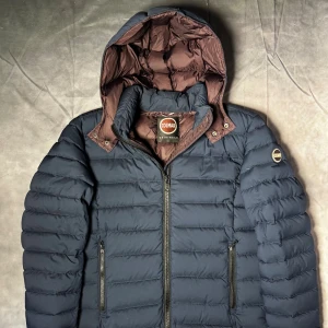 Colmar dunjacka - Säljer en mörkblå dunjacka från Colmar med brunt foder och avtagbar huva. quiltad design och Colmar-logga på ärmen. Perfekt för kyliga dagar och riktigt snygg passform. Storlek 50/M