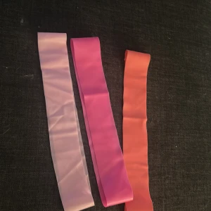 Träningsband i rosa  - Tre elastiska träningsband i olika färger: ljusrosa, rosa och orange. Perfekta för hemmaträning eller gymmet. Bandet är platt och brett, tillverkat i syntetmaterial som ger bra motstånd vid övningar. Säljs som set. Aldrig använd