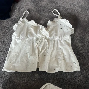 Vit linnetopp med volang och broderi - Supersöt vit linnetopp med tunna axelband, volangdetaljer vid halsen och broderade hålmönster över hela tyget. Toppen har knyt i midjan för extra passform och är tillverkad i ett lätt bomullsmaterial. Perfekt för varma dagar och en fräsch look.