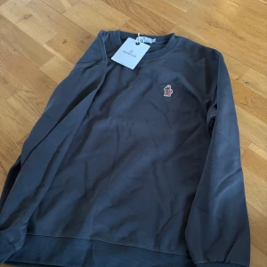 Mörkgrå sweatshirt från Moncler - Snygg mörkgrå sweatshirt från Moncler med rund halsringning och diskret broderad logga på bröstet. Tröjan har långa ärmar och ribbade muddar vid ärmslut och nederkant. Perfekt för en stilren och avslappnad look.