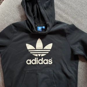 Svart Adidas hoodie med stor logga - Svart hoodie från Adidas med stor vit Trefoil-logga på bröstet. Tröjan har huva, magficka och är tillverkad i mjuk bomull. Perfekt för en avslappnad och sportig stil. Klassisk streetwear-vibe med bekväm passform och lång ärm.