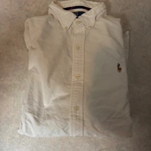 Vit slim fit skjorta från Polo Ralph Lauren - Vit skjorta från Polo Ralph Lauren i slim fit-modell, med klassisk krage och knappar framtill. Broderad logga med polospelare på bröstet. Tillverkad i mjuk bomull och har långa ärmar med manschett. Perfekt för dig som gillar stilrena och tidlösa plagg.