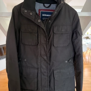 Svart parkajacka från Sebago stl S - Svart parkajacka från Sebago med fyra stora fickor framtill, hög krage och diskret logga på ärmen. Jackan har dragkedja och knappar framtill samt justerbar midja. Perfekt för kyligare dagar och har en klassisk, stilren look.