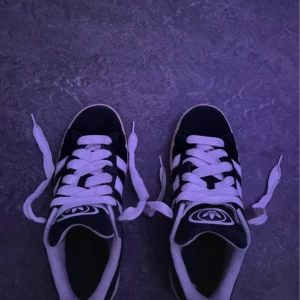 Adidas Campus svarta sneakers - Svarta Adidas Campus sneakers med klassiska vita ränder på sidorna och vit sula. Skorna har rund tå, platt sula och snörning med vita skosnören. Adidas logga på plösen. Materialet är mocka och detaljer i syntet. Perfekt för dig som gillar retrostil och streetwear.