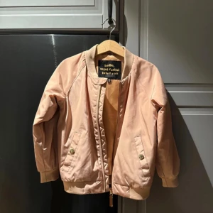 Rosa barn bomberjacka Mini Rodini med ödla - Snygg ljusrosa bomberjacka från Mini Rodini med coolt gult ödlebroderi på ryggen. Jackan har ribbade muddar, dragkedja framtill och två fickor med tryckknappar. 