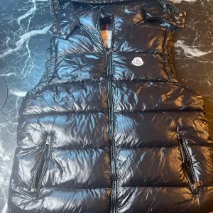 Svart dunväst från Moncler - Svart glansig dunväst från Moncler med klassisk logga på bröstet. Västen har hög krage, två dragkedjefickor och stängs med dragkedja framtill. Insidan har ett unikt Moncler-serietryck. Perfekt för lager-på-lager och streetstyle.
