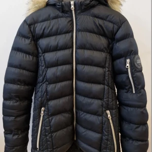 Svart pufferjacka från Vinson Polo Club - Svart pufferjacka från Vinson Polo Club med snygg fuskpäls runt huvan. Jackan har silvriga dragkedjor på fickor och ärm, quiltad design och patch på ena ärmen. Tillverkad i 100% polyamid, perfekt för kalla dagar.
