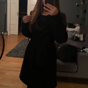 Svart lång kappa från H&M - Säljer en svart lång kappa med bälte i midjan. Klassisk och stilren modell med långa ärmar och enkel knäppning framtill. Perfekt för dig som gillar en clean och tidlös look.