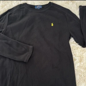 Svart långärmad tröja Polo Ralph Lauren storlek M - Svart långärmad tröja från Polo Ralph Lauren med klassisk gul broderad logga på bröstet. Tröjan är i bomull och har en enkel, stilren design med rund hals. Perfekt för en clean och avslappnad look.
