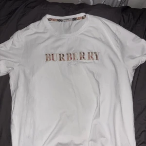 Vit Burberry t-shirt - Vit t-shirt från Burberry i storlek M med klassisk Burberry-logga broderad på bröstet och rutigt band på insidan av kragen. T-shirten har rund hals och korta ärmar, tillverkad i mjuk bomull för skön känsla. Perfekt för dig som gillar stilrena designer med lyxig touch. Inte äkta